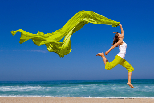 Woman running and jumping on sand shoreline of beach with green scarf.